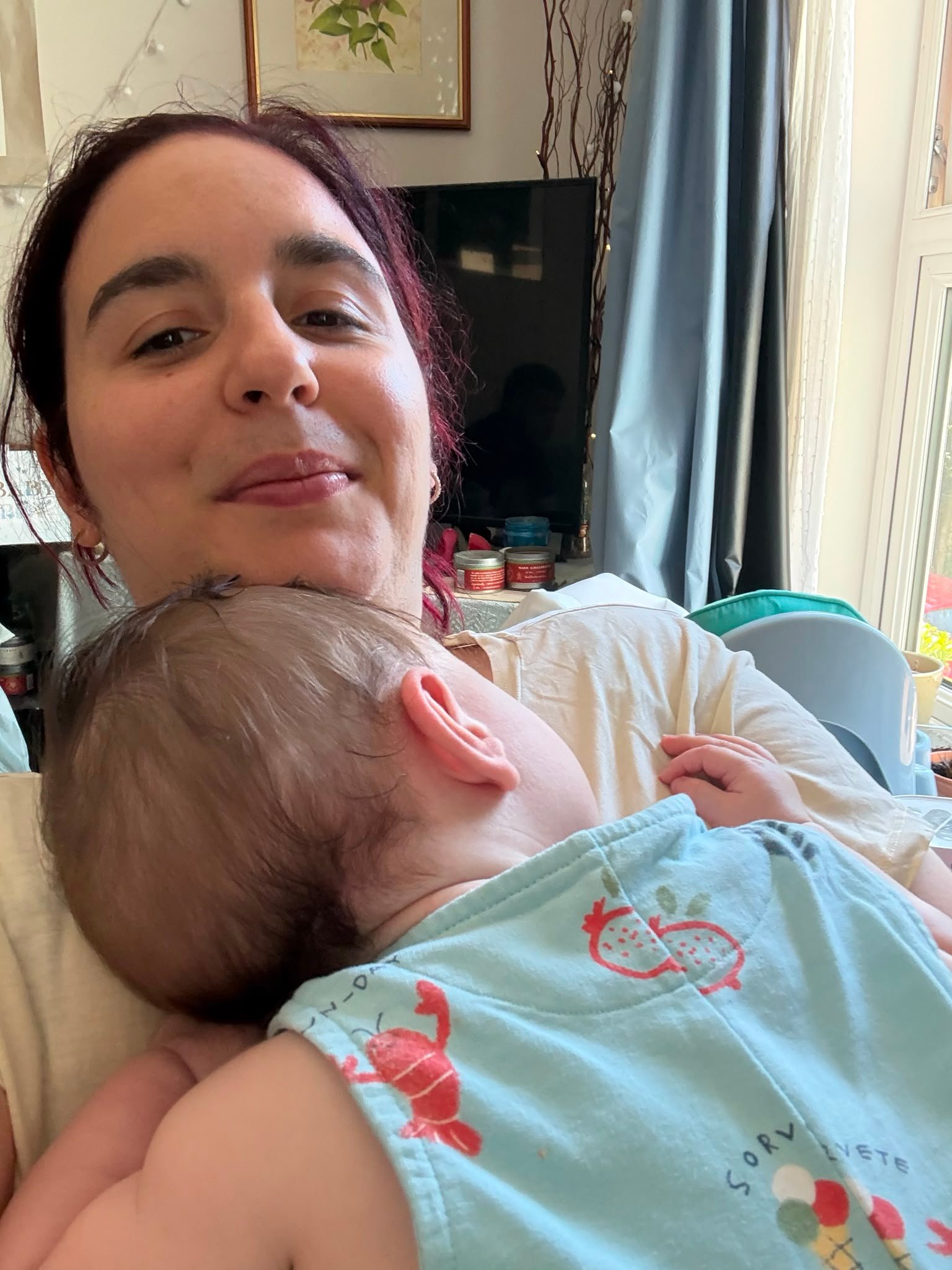 A mother smiling softly at the camera while her baby rests against her chest at home, with a TV and window in the background.