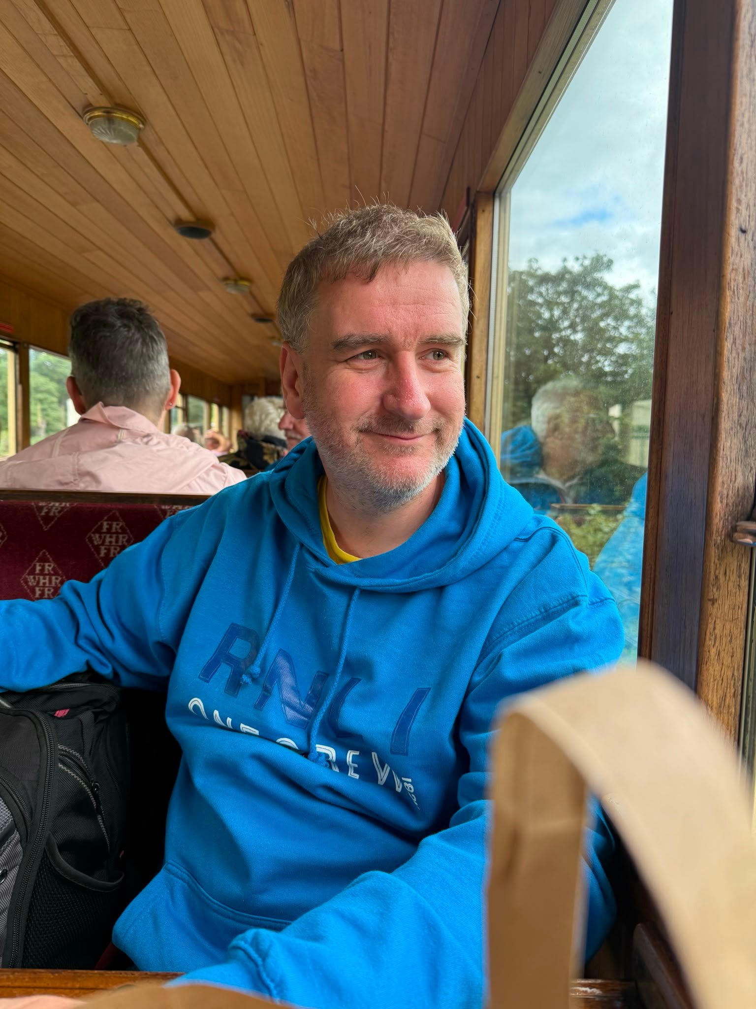 James wearing a blue RNLI hoodie, smiling slightly, looking out of a train window.