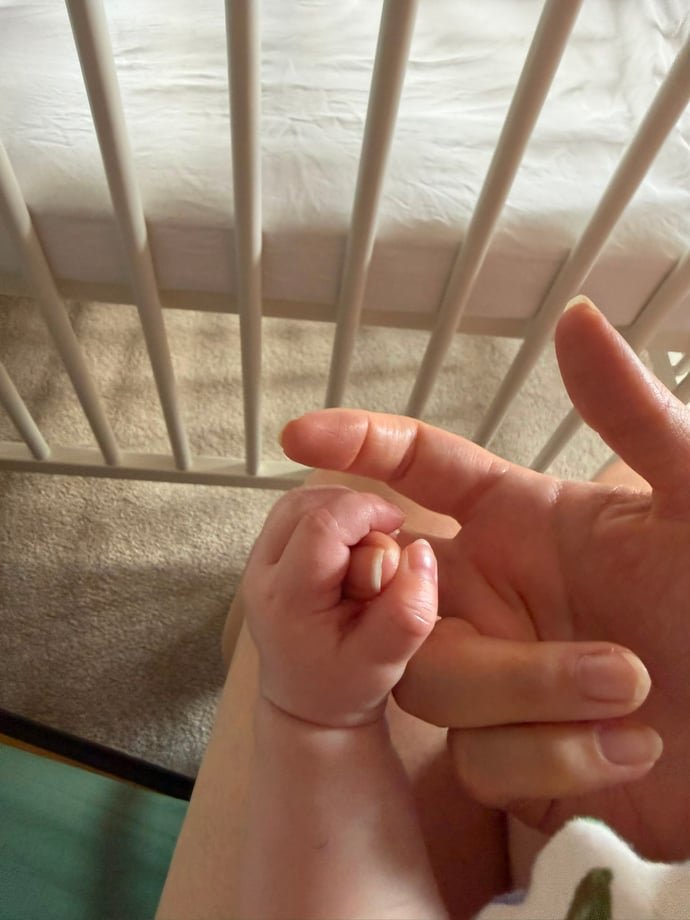 Baby holding an adult’s finger beside a cot