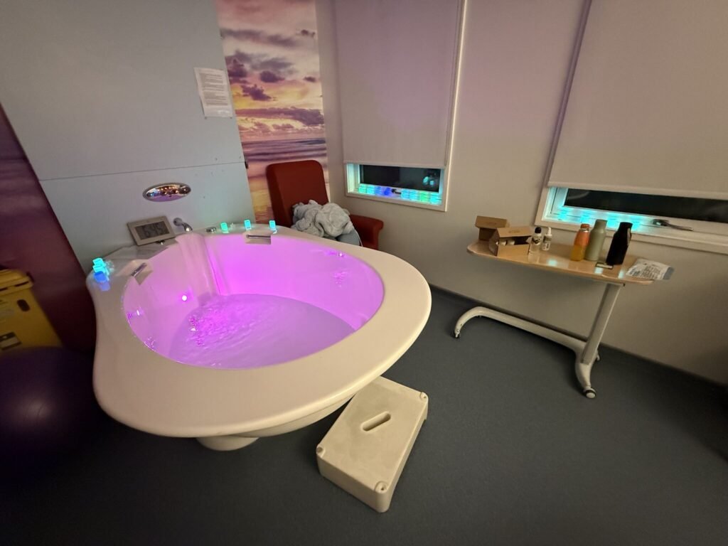 A hospital birthing pool filled with water and illuminated with pink and purple lights, in a dimly lit maternity room with blinds drawn and a chair beside the pool.