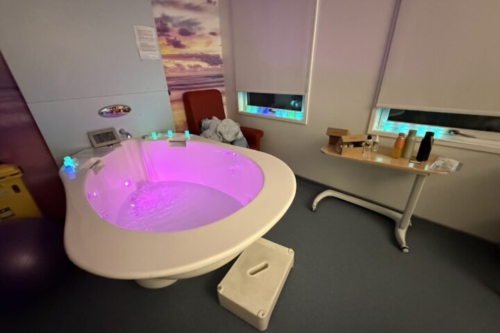 A hospital birthing pool filled with water and illuminated with pink and purple lights, in a dimly lit maternity room with blinds drawn and a chair beside the pool.