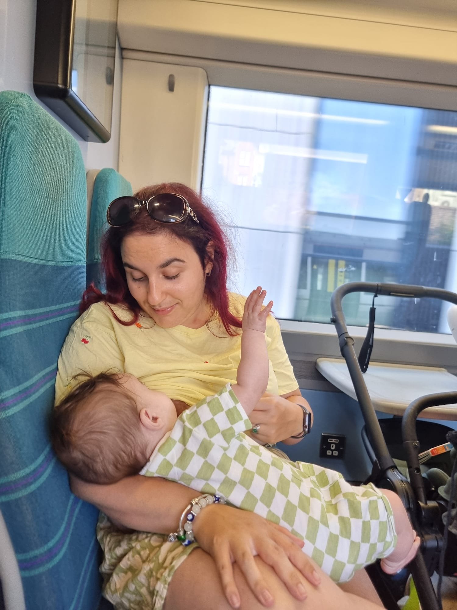 A woman sitting on a train holding a baby across her lap. The baby’s face is turned away from the camera, with one arm raised. A buggy is next to them by the window.