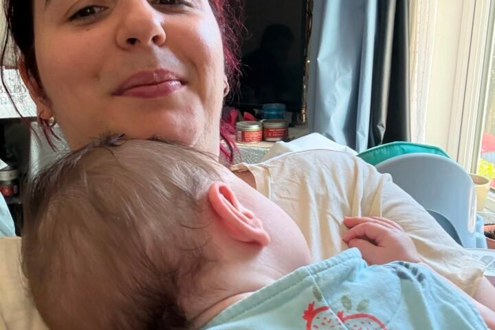 A mother smiling softly at the camera while her baby rests against her chest at home, with a TV and window in the background.
