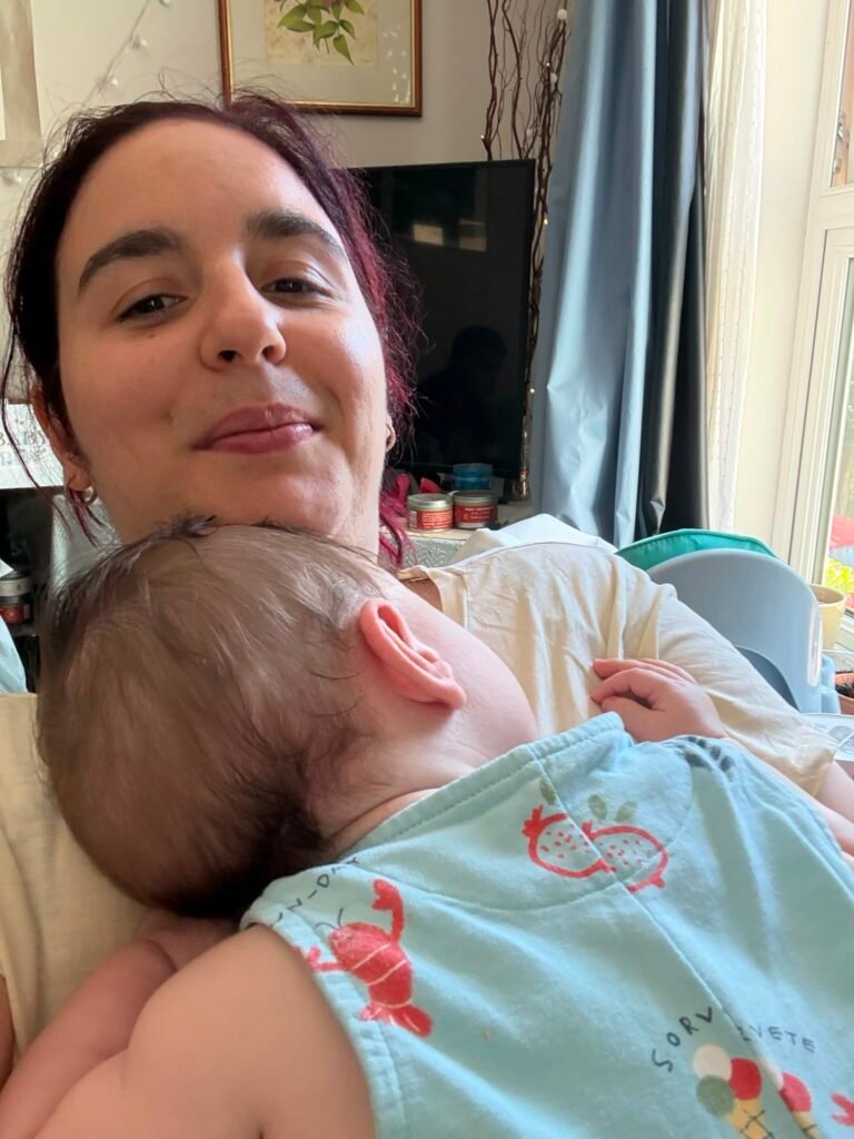 A mother smiling softly at the camera while her baby rests against her chest at home, with a TV and window in the background.